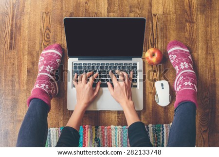 Workspace in home: fingers on laptop keyboard, red apple, legs in socks on a wooden floor and colored carpet