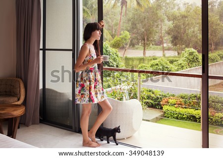 Beautiful Young Woman near big Window with black cat  holding cup and relax inside home