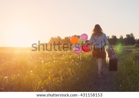 Young Woman walking with big suitcase and colored balloons on meadow road. Sunrise, wild flowers, summer  freedom and travel