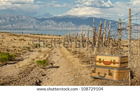 Vintage luggage sitting by a dirt road with mountains and lake