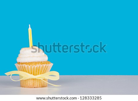 Single vanilla cupcake with yellow candle, bow and sprinkles on a blue background and white table