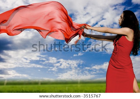 The girl in a red dress and with a red shawl flying on a wind on a green floor on a background of the blue sky with clouds