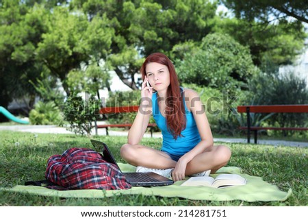 student in park with the book and computer talking on cell phone