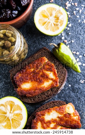 Rye bread sandwiches with fried halloumi cheese, sun-dried olives in bowl, lemon, capers and pepper on gray stone background