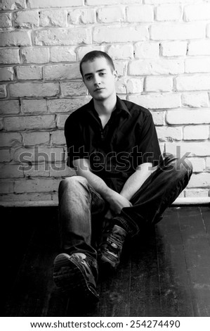 Handsome pensive young man sitting on the floor near the white brick wall, toned black and white