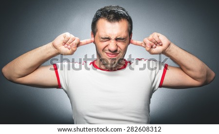 Noise - loud. Closeup portrait of a young angry unhappy stressed man covering his ears closing eyes over gray dark background