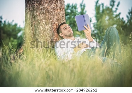 Young man read book and repose in grass. Guy leaning against a tree relaxing - resting
