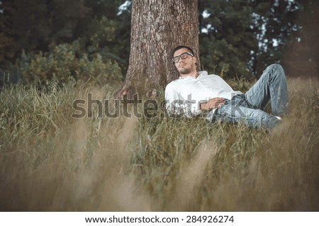 Young handsome man repose on grass against a tree. Gorgeous guy lie down on meadow in nature. Outdoors - outside