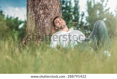 Young handsome man repose on grass under a tree. Gorgeous guy lie down on meadow in nature. Outdoors - outside