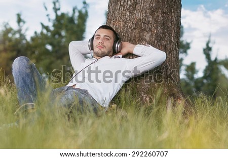 Young man listen music - enjoy - repose in grass. Guy leaning against a tree relaxing - resting. Outdoors - outside