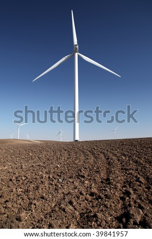 White power generating windmills under blue sky and recently tilled agricultural land.