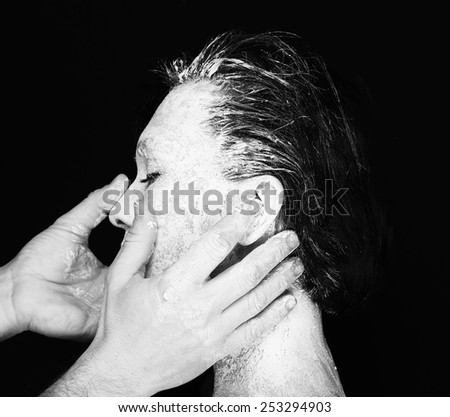 Black and white portrait of human hands working with woman with clay on face on black background