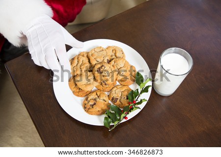 A view from above a plate of cookies and glass of milk as the white gloved hand of Santa Claus reaches for one