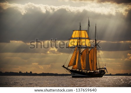 A three-master sailing on the Elbe near Hamburg. The evening light breaks through the gaps in the clouds behind the ship, illuminating the sails from behind. Processed to give it an antique look.