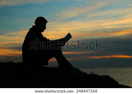 Silhouette of a man sitting on breakwater in evening near sea, reads book