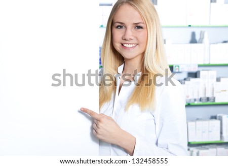 Attractive saleswoman in pharmacy points at blank white sign.