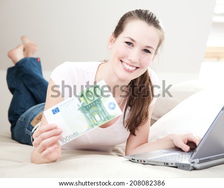 Attractive young woman on her bed with laptop holds 100 euro.