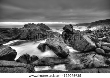 A black and white stormy seascape with big rocks and moving sea water. Dark rain clouds hanging in the sky.