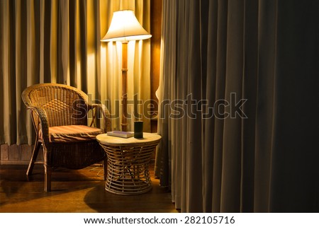 Rattan chair and stool under the light from the lamp with bamboo cup and the book surround by beautiful curtain.