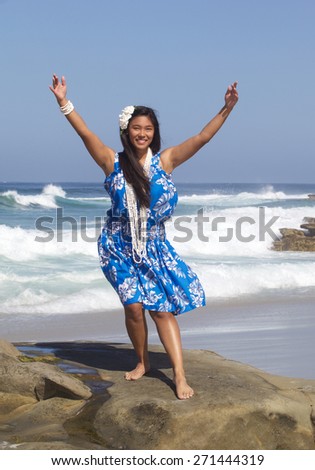 A beautiful teenage girl dancing hula on an empty beach.  She is wearing a traditional hula dance outfit, a long shell necklace and a plumeria flower in her hand.