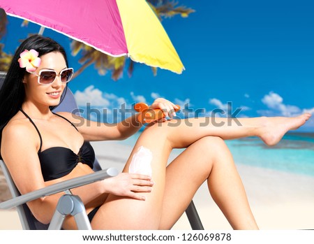 Beautiful happy woman in black bikini applying sun block cream on the tanned body.  Girl  holding orange sun tan lotion bottle.