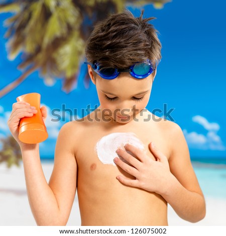 Schoolchild boy applying sun block cream on the tanned body.  Boy holding orange sun tan lotion bottle.