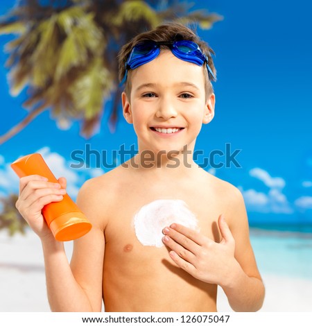 Happy schoolchild boy applying sun block cream on the tanned body.  Boy holding orange sun tan lotion bottle.