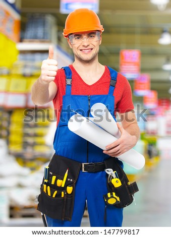 Portrait of smiling handyman with tools and paper showing thumbs up sign  stands at warehouse