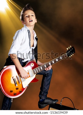 A young white boy sings and plays on the electric guitar on the stage with bright lights