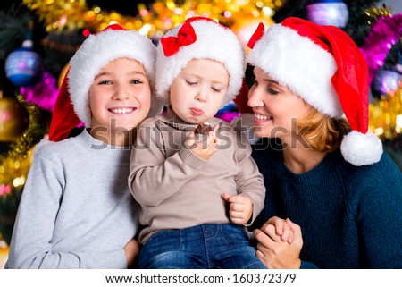 Toddler eats the sweet candy in a circle of family. Smiling young mother with two sons in Santa hats.