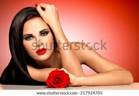 Portrait of a beautiful woman with red lips holds the rose in hand .   Fashion model with long hairs posing at studio over red background