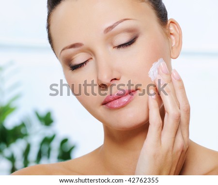 Candid woman face with closed eyes applying  moisturizer cream on her cheek