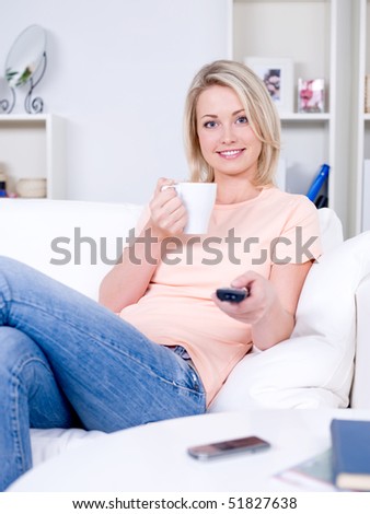 Smiling young woman with cup of coffee sitting on the sofa and switching the TV channels. Holding the  remote control