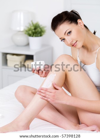 Beautiful young smiling woman sitting on a bed and applying cream on her attractive legs - vertical