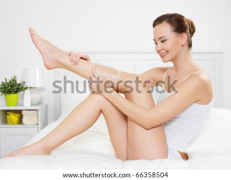 Beautiful young woman depilating skin on legs by waxing is in the bedroom - indoors