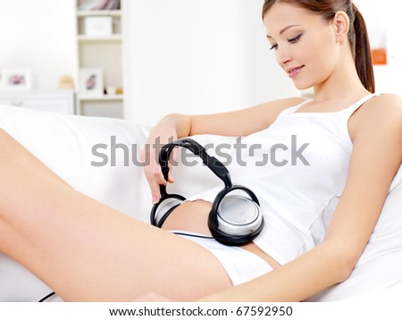 Pregnant woman sitting on white sofa at home and listening music in headphones