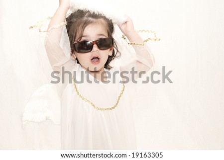 cute toddler girl dressed in angel costume wearing sunglasses taking off her halo against white background