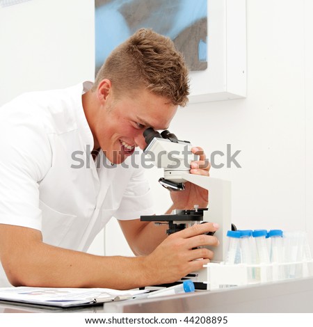 A young male lab technician looks at slides through a microscope whilst comparing the results to earlier tests.