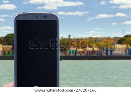 Smatrphone and a landscape. Idea for travel apps, digital detox, taking shots, accessing apps, Internet, blogs and others. The blur image are Colored houses at Porto Seguro coast - Bahia - Brazil