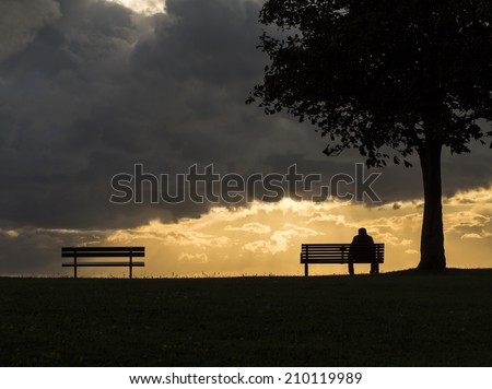 Silhouette portrait of a lonely anonymous man sitting on a bench as dark clouds above him roll, with the sun beginning to set behind