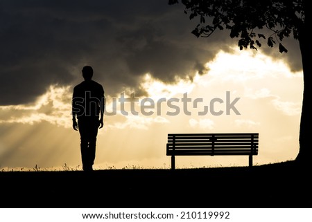 Silhouette portrait of an anonymous man walking away from a bench as dark clouds roll above him. The sun behind creating light between the clouds