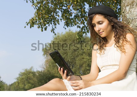 An attractive young woman reading a tablet on a bright sunny day whilst under a tree