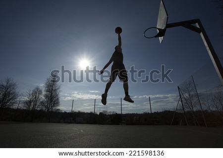 Silhouette of a basketball player in mid air on an outdoor basketball court about to slam dunk on a bright sunny day, motion blur