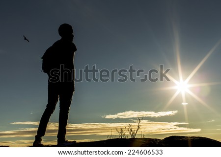 Silhouette male with a backpack on top of a mountain or hill looking into the distance as the sun begins to set