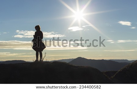 Anonymous silhouette female runner standing on a mountain top and checking her playlist on her music device as the sun flares in the background