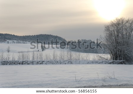 Partially clouded sky with the Sun shining through it, illuminating snow covered fields, and a traditional Norwegian slanted fence.