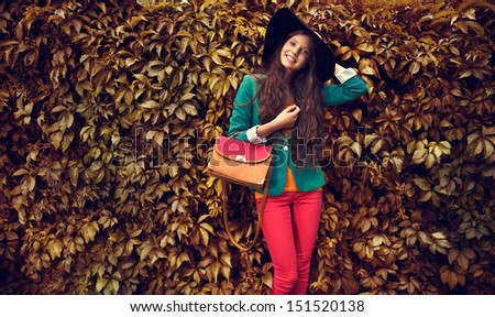 Beautiful autumn woman holding handbag wearing black hat against leaves