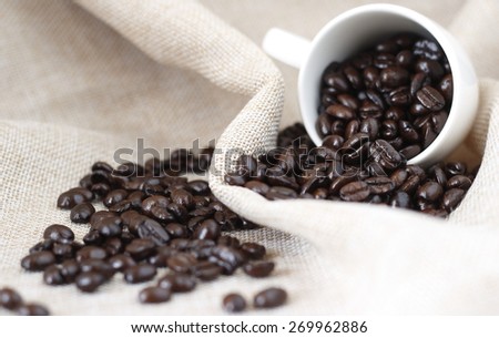 coffee bean flowing from white coffee cup on bleached jute cloth (selective focus)