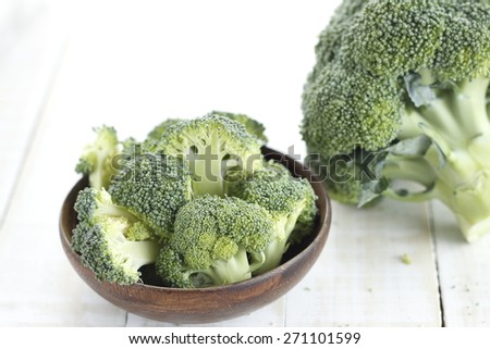 pieces of broccoli in wooden bowl and broccoli on white wooden background