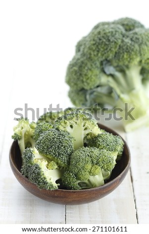 pieces of broccoli in wooden bowl and broccoli on white wooden background